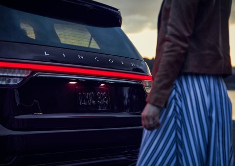 A person is shown near the rear of a 2024 Lincoln Aviator® SUV as the Lincoln Embrace illuminates the rear lights | Jenkins & Wynne Lincoln in Clarksville TN